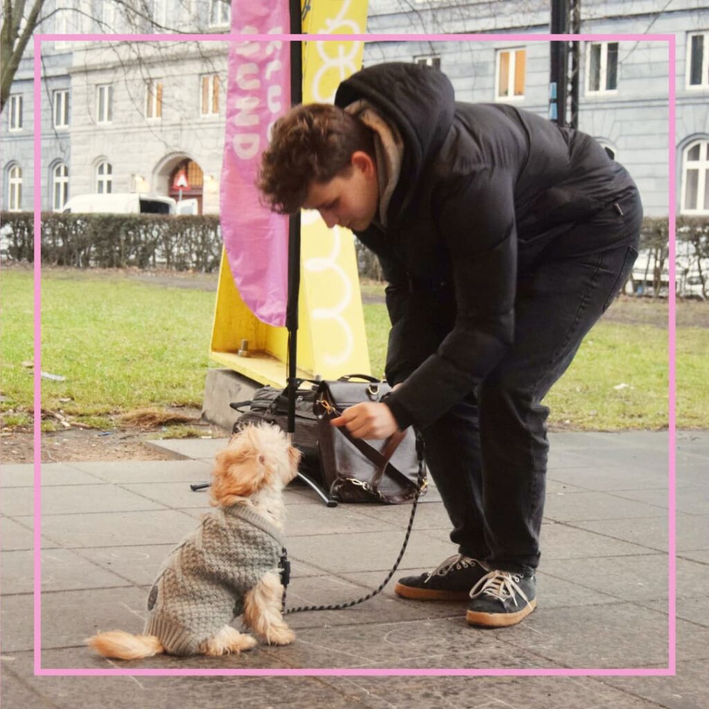 En mand i sort jakke og sneakers bukker sig ned og giver godbid til en lille lys hund iført grå striktrøje, der sidder pænt på fortovet i snor foran et lyserødt Klub Hund-banner.