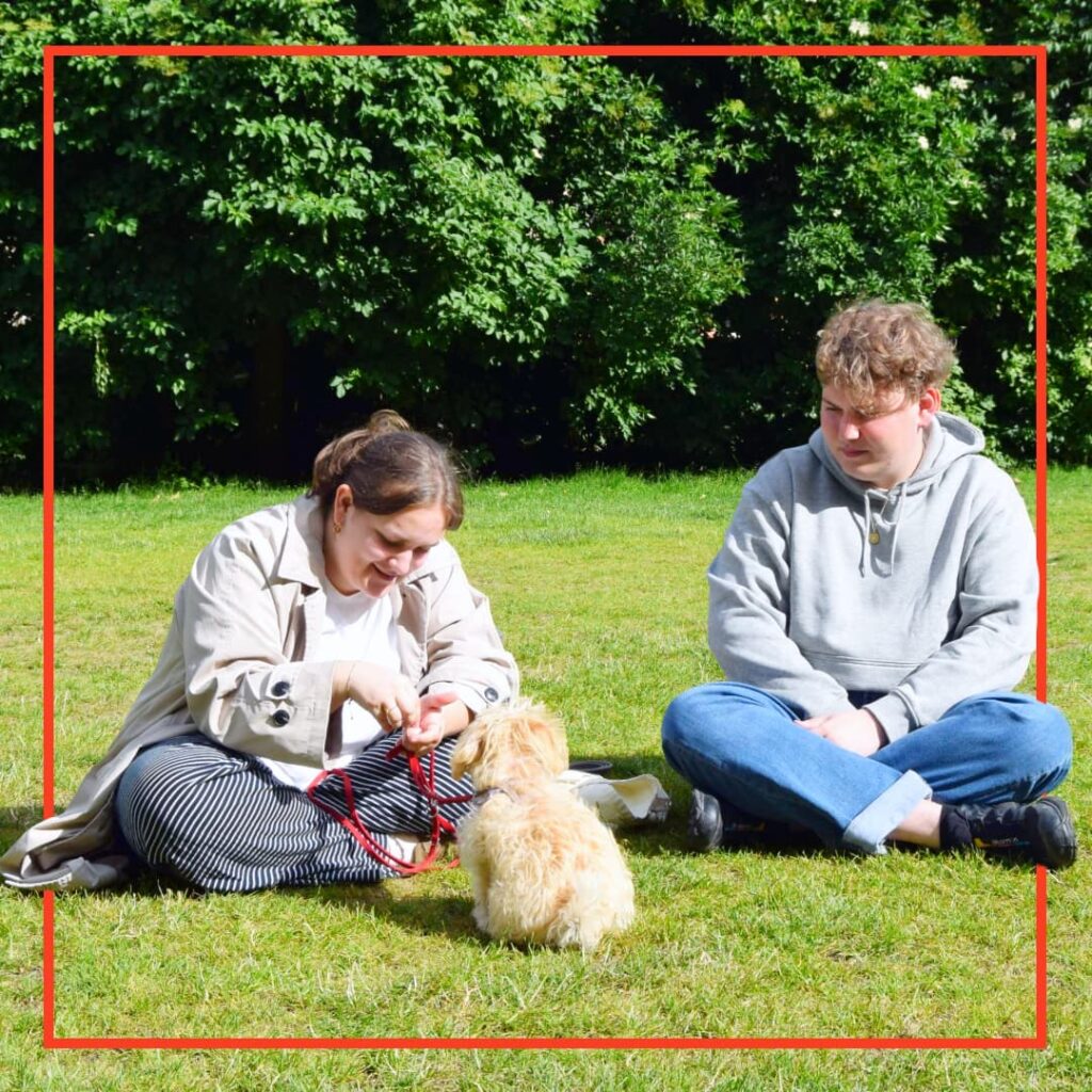 To hundeejere fra Klub Hund sidder på græsset i parken, mens de træner en lille lys hvalp med snor.