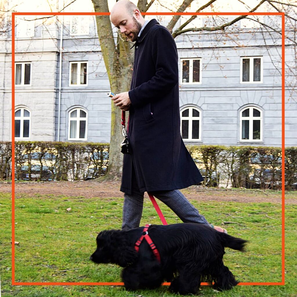 En mand går tur i en park med en sort hund i rødt sele. Manden smiler ned mod sin hund, mens hunden snuser i græsset.