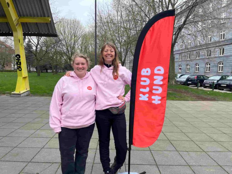 Michelle og Marie fra Klub Hund står smilende i lyserøde trøjer ved et Klub Hund-banner.