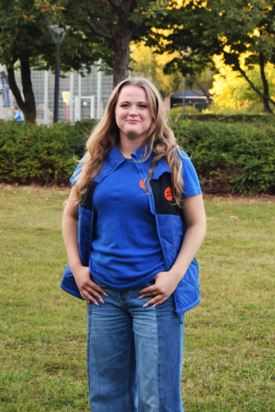 Hundetræner Signe fra Klub Hund iført blå træningstøj, stående udendørs på en græsplæne.